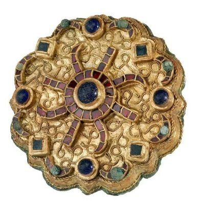 The brooch has the form of a box, its basic shape is round, with delicate indentations that form a kind of lobes. In the center is a blue round cabochon surrounded by a frame of flat garnets. Eight rays made of flat garnets, four straight and four curved rays emerge from the frame. The edges of the brooch are decorated with a repeating pattern of blue and green cabochons, and crescents made of flat garnets with a small green cabochon in the middle.
All the setting of stones and glass are raised to the surface of the brooch. The background is a golden sheet decorated with irregularly winding pieces of wire.